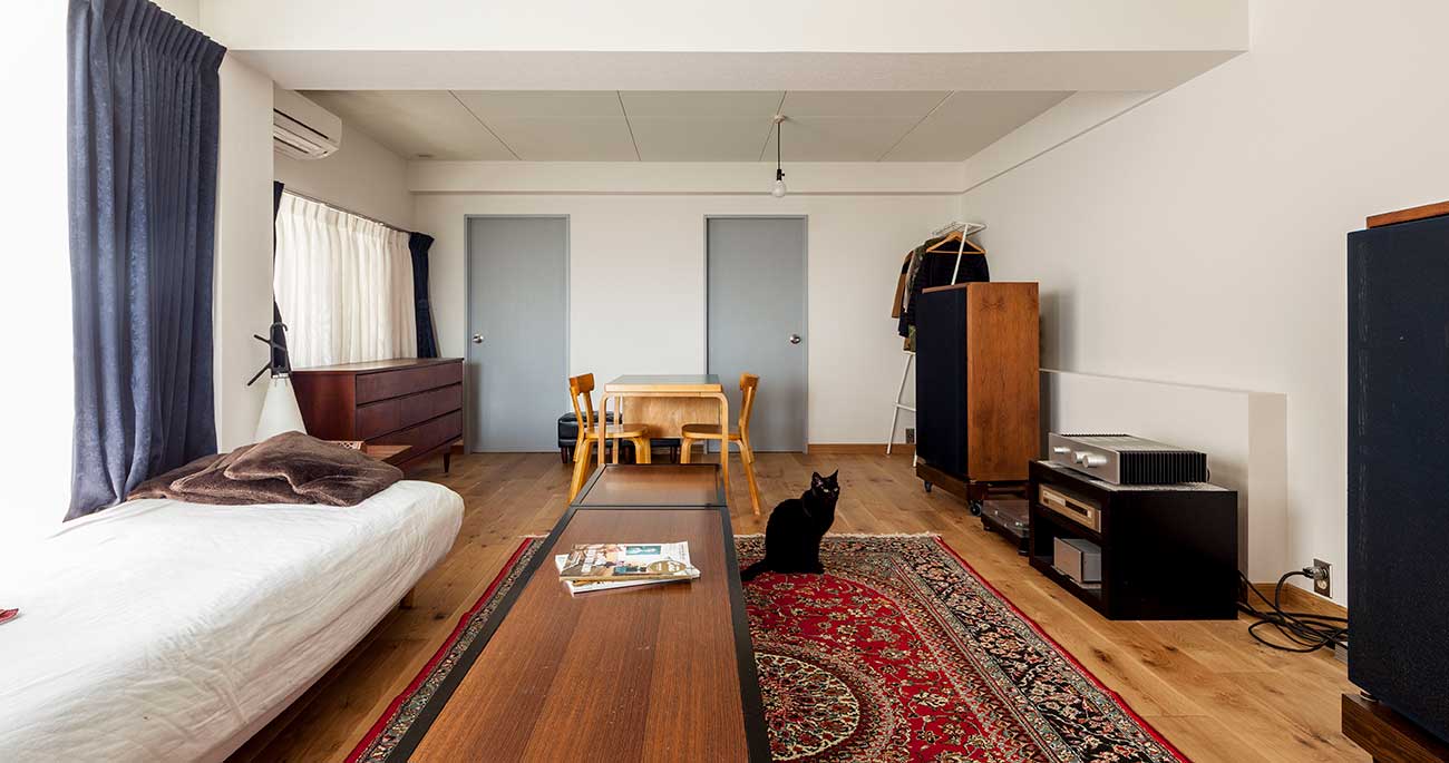 リビングの風景。白い天井と壁、幅広のフローリングがナチュラルでシンプルな空間。奥の壁には2つのドアが並び、木のダイニングセットやチェスト、アイアンと木でできた横長のテーブルや、立派なオーディオ機器があり、ペルシア絨毯の上には飼われている黒猫が座っている