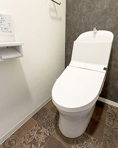 トイレ空間。コンパクトな便器の背面は濃いグレーの壁。側面は白い壁になり、床は洗面脱衣室とお揃いのこげ茶色の模様のある床です。