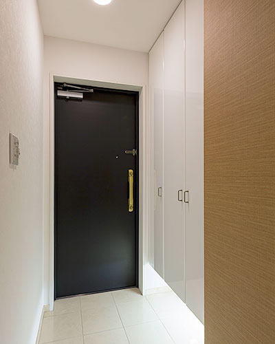 白い天井と壁、土間部分には白いタイルが貼られた玄関スペース。ドアは黒く手摺りはゴールド。白い造り付けの玄関収納があり、足元が間接照明で照らされている。