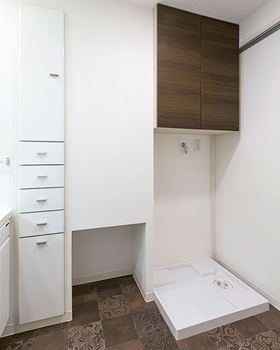 洗面脱衣室には壁と色を合わせた白い扉の天井まである収納と洗濯機置場があります。白い壁と天井の空間に、ランダムに模様の描かれたこげ茶色の床が対照的です。