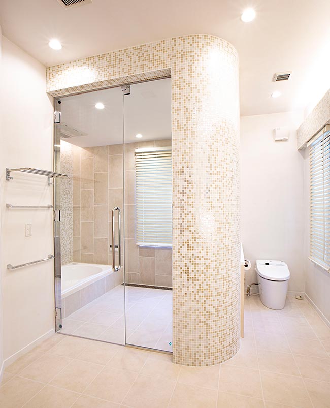 白を基調とした水廻り空間。ガラスの扉と曲線のタイルの壁で仕切られた浴室空間と、その横にあるトイレ