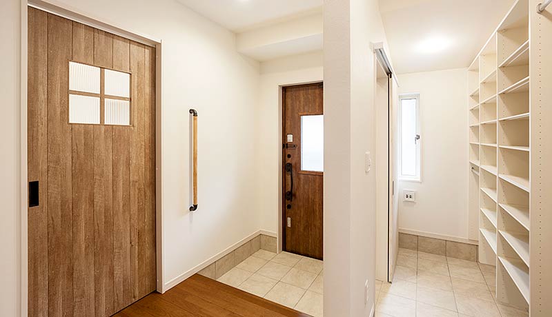 玄関スペースの写真。正面に茶色の木目調の玄関ドアがあり、土間続きの右側には一面が棚になったウォークインシューズクローゼットがある