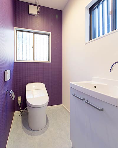トイレ空間。左側と奥の壁はビビットな紫色のクロス、右側の壁は白いクロスが貼られ白いタンクレスの便器が設置されている。正面と右側の壁には腰窓があり、右側には白い洗面台がある