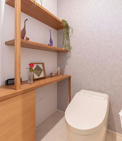 トイレ空間の様子。タンクレス便器の背面には明るいグレーの壁。左側の白い壁には高さのある木の棚が設置され、小物やグリーンが飾られている。