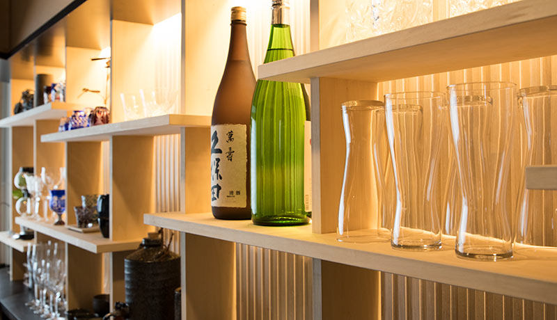 ランダムな高さの木の棚に、日本酒や様々なグラスが飾られ間接照明に照らされている