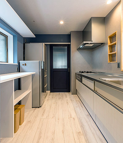 キッチンスペースの様子。右側にはシステムキッチンがあり、左側には冷蔵庫と棚が並んでいる。正面の奥の壁には洗面へと続く青いドアがある。