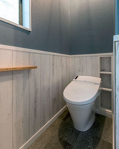 トイレ。青い壁には白い腰壁があり、床はグレーの木目調。白いタンクレスの便器が設置されている。